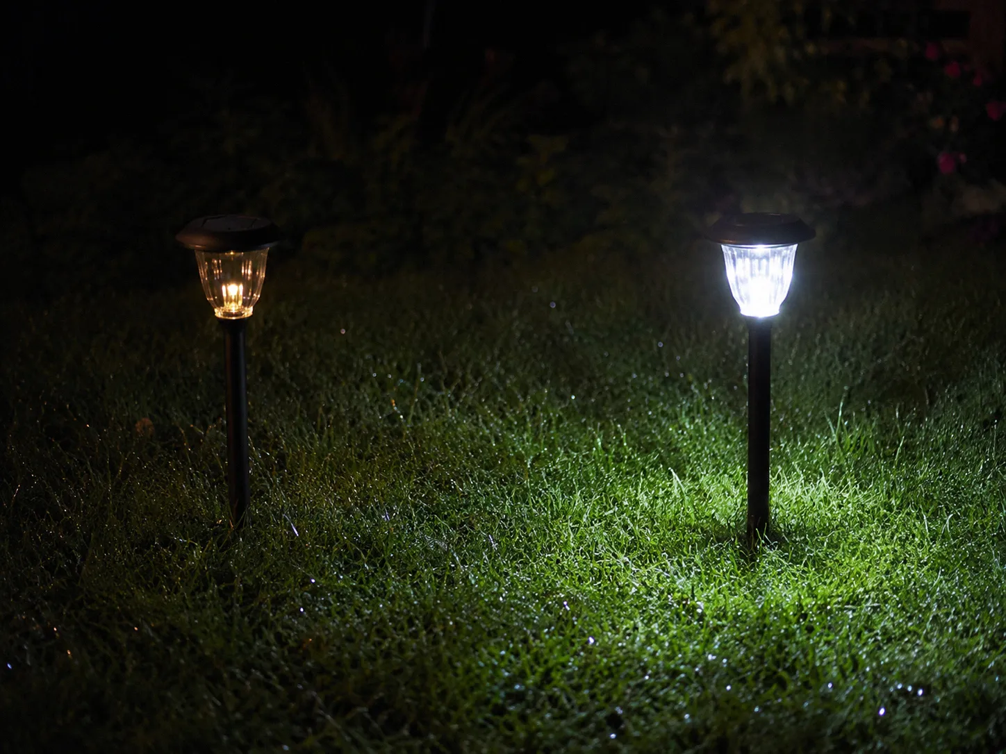 Two identical solar garden stake lights at night, the left one dim and flickering with a faint orange glow, the right one at full brightness casting crisp white light on the dewy grass