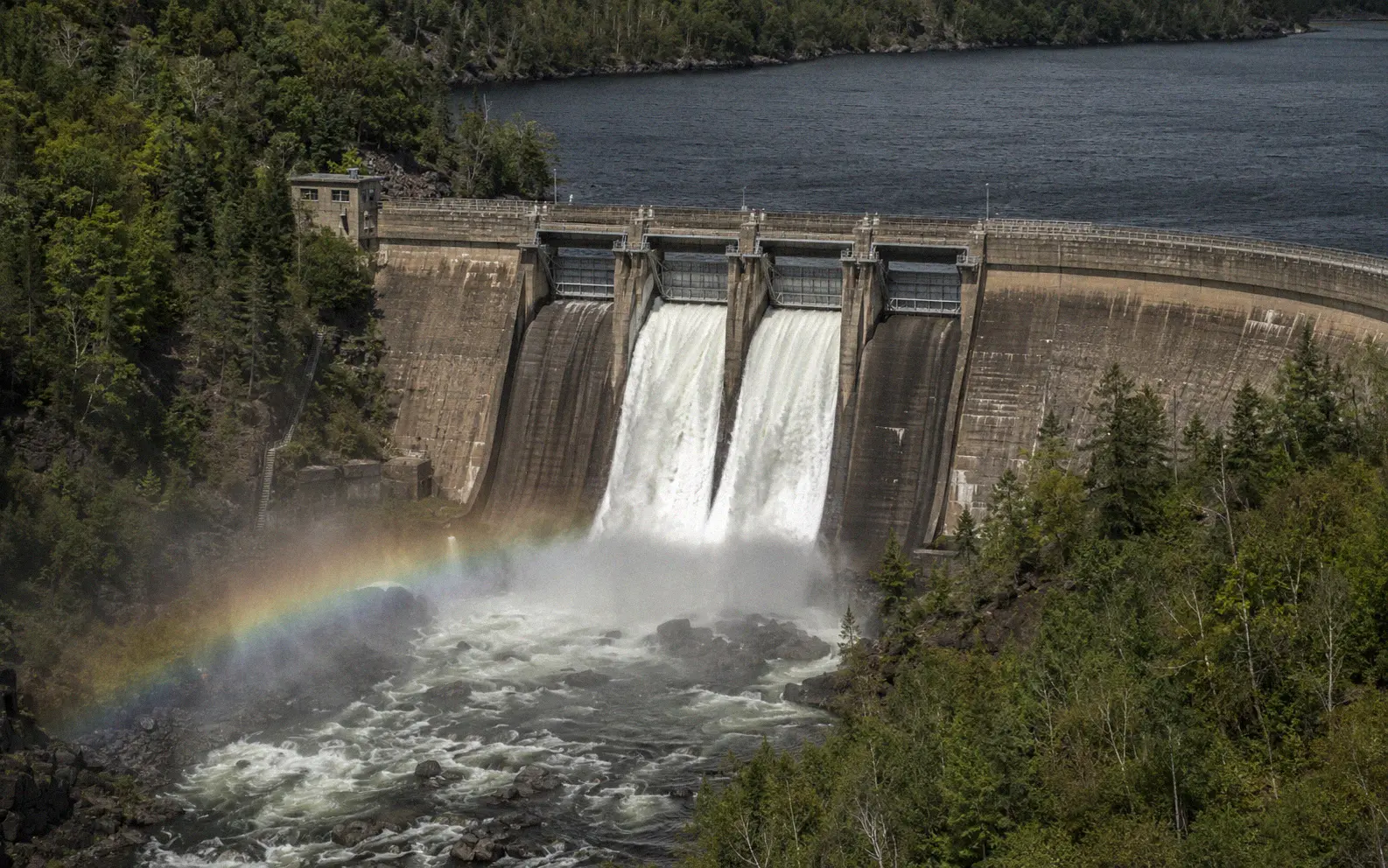 Large concrete hydroelectric dam releasing powerful white water through spillways with a faint rainbow in the mist and forested hillsides on both banks