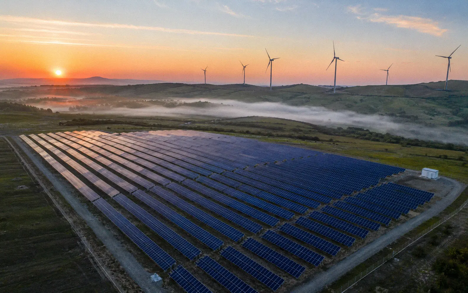 Aerial drone view of a large solar farm at sunrise with morning dew on panels and wind turbines on surrounding green hills